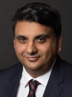 A headshot of Jadbinder Seehra in a suit smiling in front of a dark background