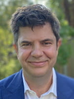 A headshot of Paul Ashley wearing a blue suit, he is in front of a green leafy background